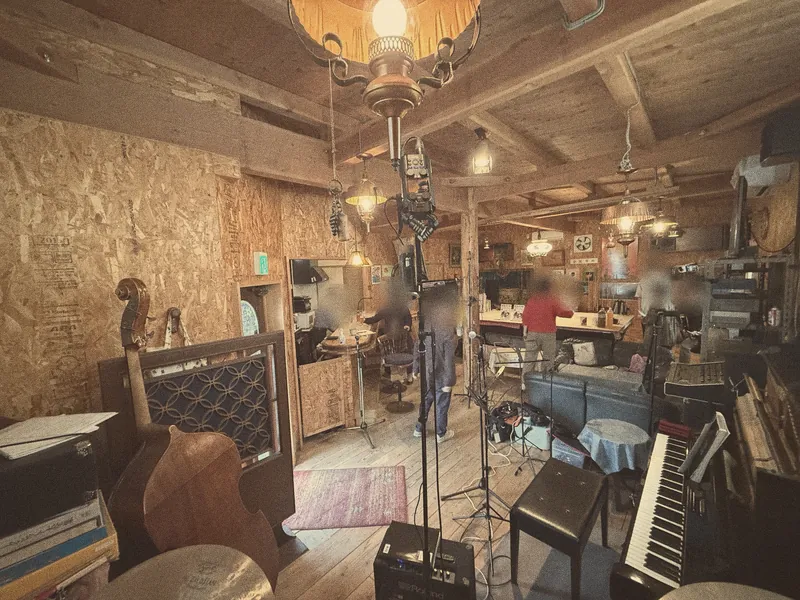 The room seen from the drum kit: a double bass and piano in front, the counter and tables behind, lamps hanging from wooden beams